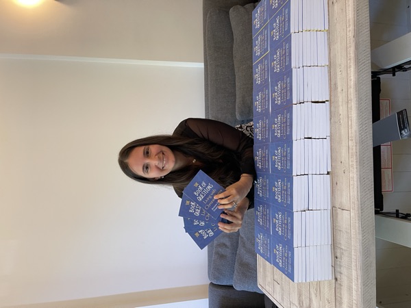 Brianna today, with stacks of her book The Book of Questions: Living with Chronic Illness